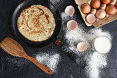 Hot pancake in black pan on black table with flour, milk and eggs