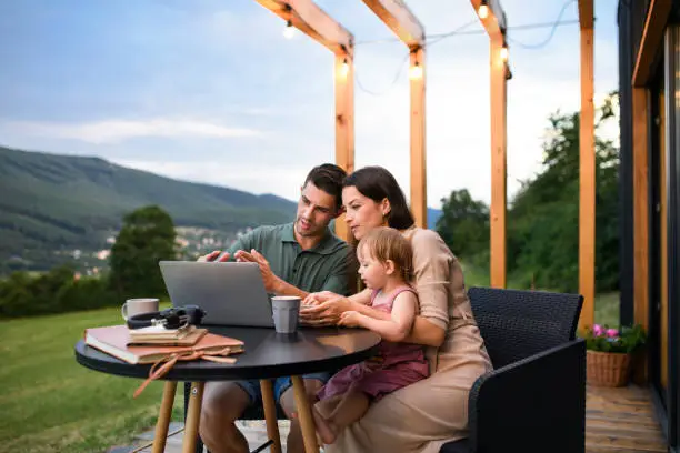 Young family with small daughter using laptop outdoors, weekend away in container house in countryside.
