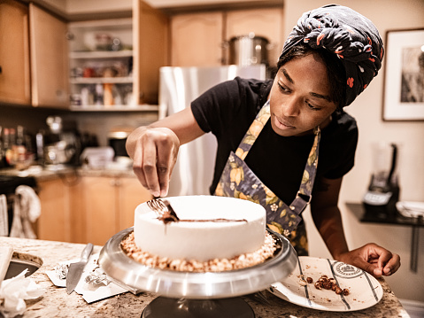 Young black woman baking a sweet cake from the scratch in her kitchen. She is putting icing on the new cake.  Interior of private home.