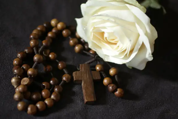 Wooden rosary with Jesus Christ holy cross crucifix and white rose on black background. Believe in God concept. The symbol of Catholic meditation prayer.