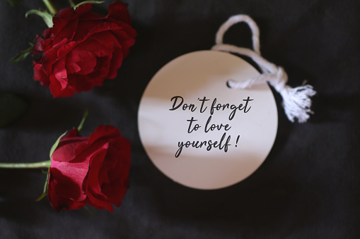 Motivational text message on white tag label paper - Do not forget to love yourself. With red roses on dark black background. Self love and care concept.
