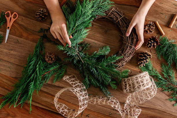 Overhead shot of hands making handmade Christmas wreath with twigs, pine cones, cinnamon sticks, yew and thuja branches and natural string. DIY home decor. stock photo