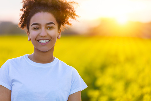 Beautiful smiling mixed race African American girl teenager female young woman with perfect teeth at sunset or sunrise in a field of yellow flowers