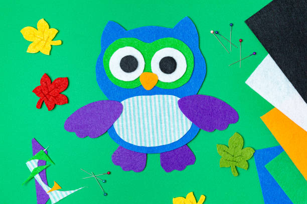 Craft, applique of an owl from multi-colored felt on a green background. Creative activities at home with children. Sewing soft toys, hobby. Workplace. Felt bird, owl, big eyes. Flat lay, top view.