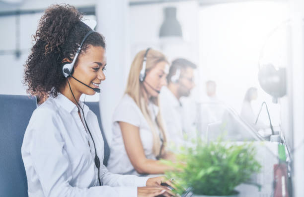 Call center worker accompanied by her team. Call center worker accompanied by her team. Smiling customer support operator at work. Young employee working with a headset. service stock pictures, royalty-free photos & images