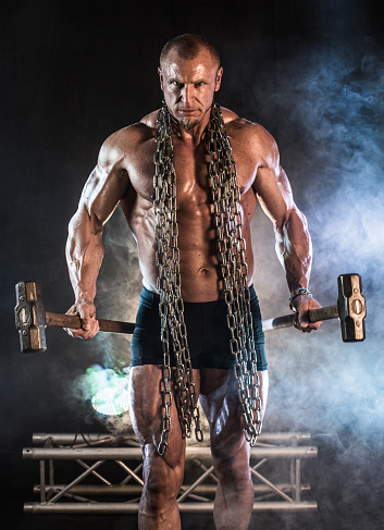 A middle aged ripped bodybuilder walking towards camera looking mean holding a sledgehammer in each hand and heavy chains around his neck.  Dramatic lighting and smoke behind him.