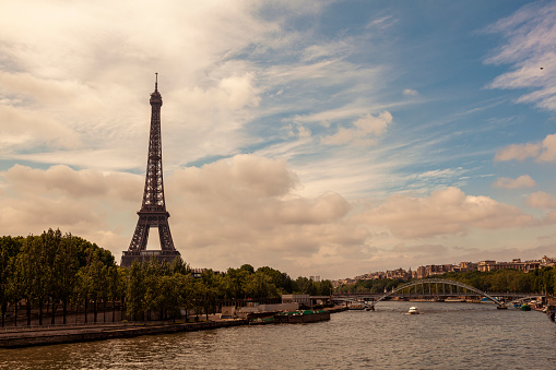 scenic view of Paris, France in spring with Seine river on one side and the famous Eiffel tower on the other. A small metal pedestrian bridge, trees, a river boat, buildings are seen in background.