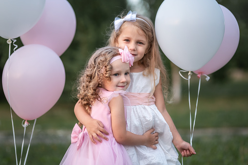 Two little girls in pink and white dressses with balloons hugging in the park