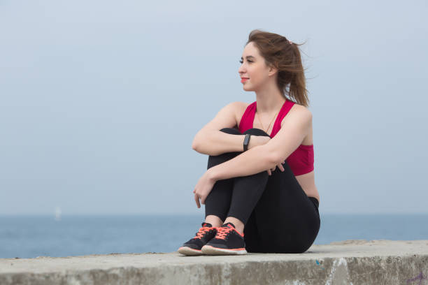 Girl sitting with her knees to chest outdoors Young woman in black leggings and red tank top hugging her knees on the pier hugging knees stock pictures, royalty-free photos & images