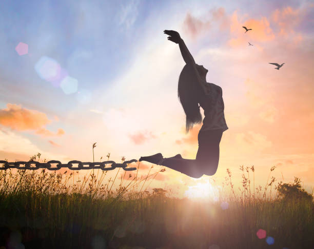 International day for the remembrance of the slave trade and its abolition concept Silhouette of a woman jumping with her hands raised and broken chains at meadow autumn sunset releasing stock pictures, royalty-free photos & images