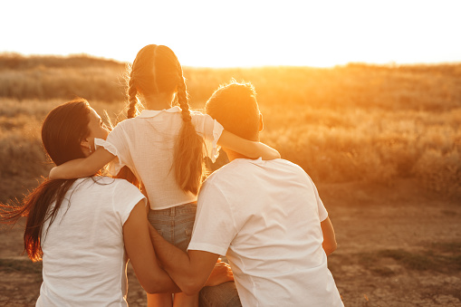 Back view of parents holding and supporting little girl while standing in field and looking at setting sun in nature