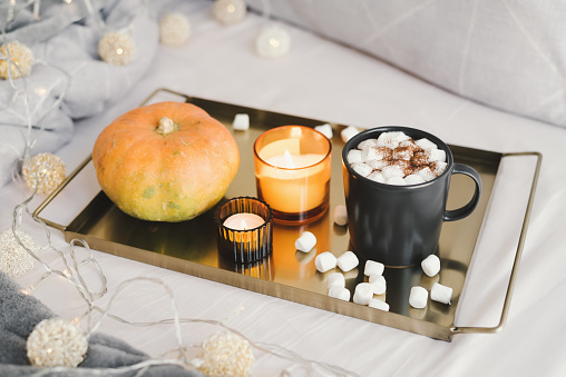 Hot chocolate with marshmallow and cocoa powder in a black mug, burning candles and pumpkin on a metal tray served in bed. Fall season inspired dessert
