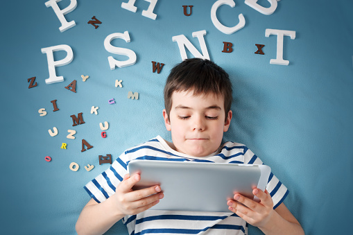 Seven years old child is lying with tablet and letters on blue background.