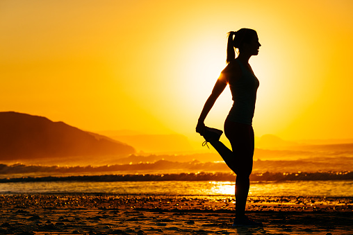 Fitness woman exercising and stretching legs on beautiful summer sunset or morning at beach. Female runner warming up or cool down.