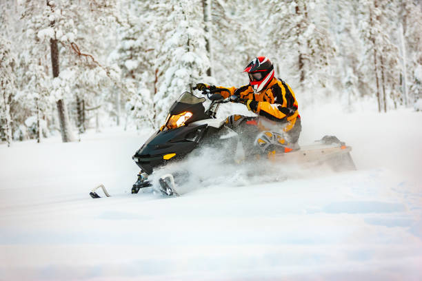 humano en el atuendo de un corredor en un mono multicolor y un casco, conduciendo una moto de nieve por la superficie de nieve profunda en el fondo del bosque nevado. - motoesquí fotografías e imágenes de stock