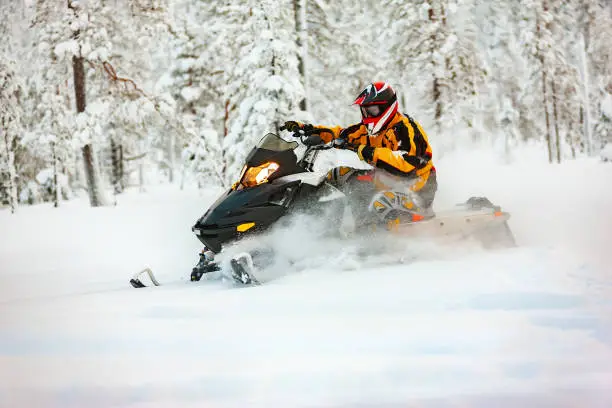 A man in the outfit of a racer in a yellow-black overalls and a red-black helmet, driving a snowmobile at high speed riding through deep snow against the background of a snowy forest.