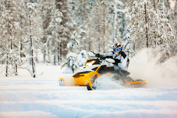 racer en el conjunto de un mono y un casco, conduciendo una moto de nieve por la superficie de nieve profunda en el fondo del bosque nevado. - motoesquí fotografías e imágenes de stock