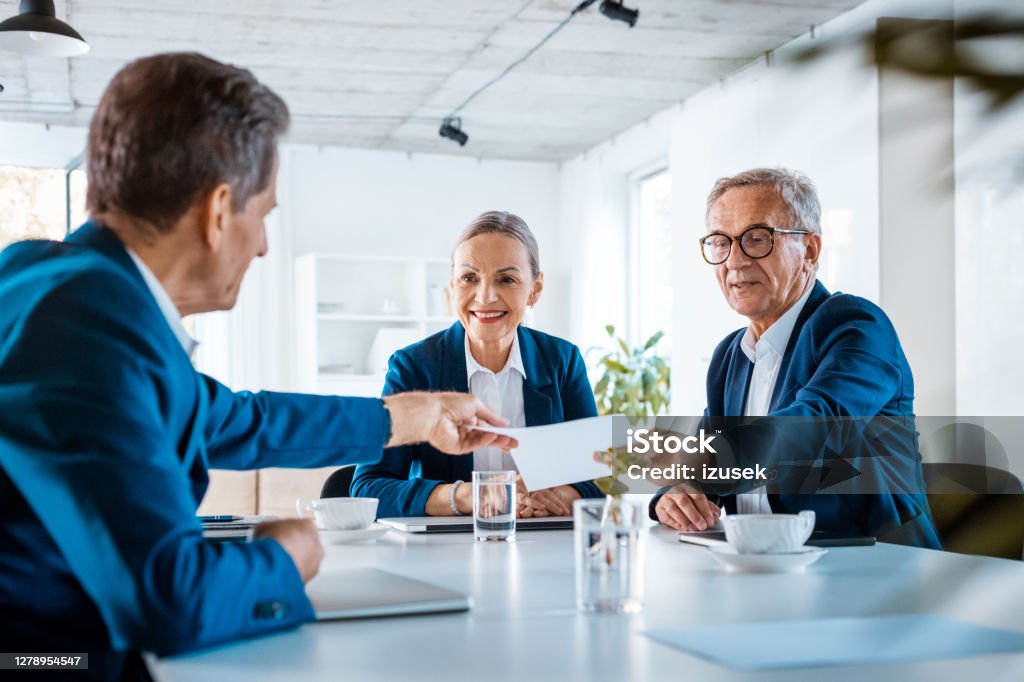 Senior business people during meeting Senior businesswoman and businessmen wearing elegant suits sitting at the table in the office and discussing during business meeting, exchanging contract. Exchanging Stock Photo
