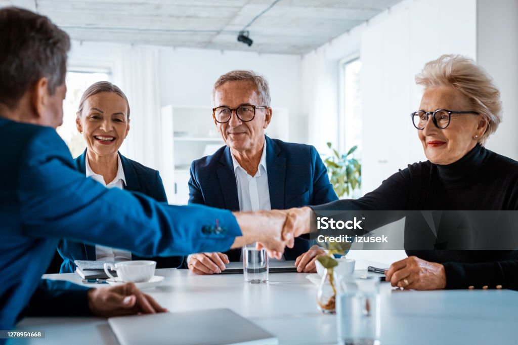 Senior business people during meeting Senior businesswoman and businessmen wearing elegant suits sitting at the table in the office and discussing during business meeting, shaking hands. Navy Blue Stock Photo