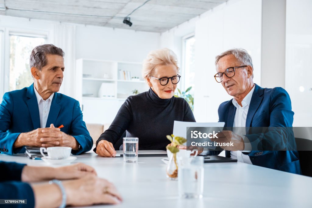Senior business people during meeting Senior businesswomen and businessmen wearing elegant suits sitting at the table in the office and discussing contract during business meeting. 65-69 Years Stock Photo