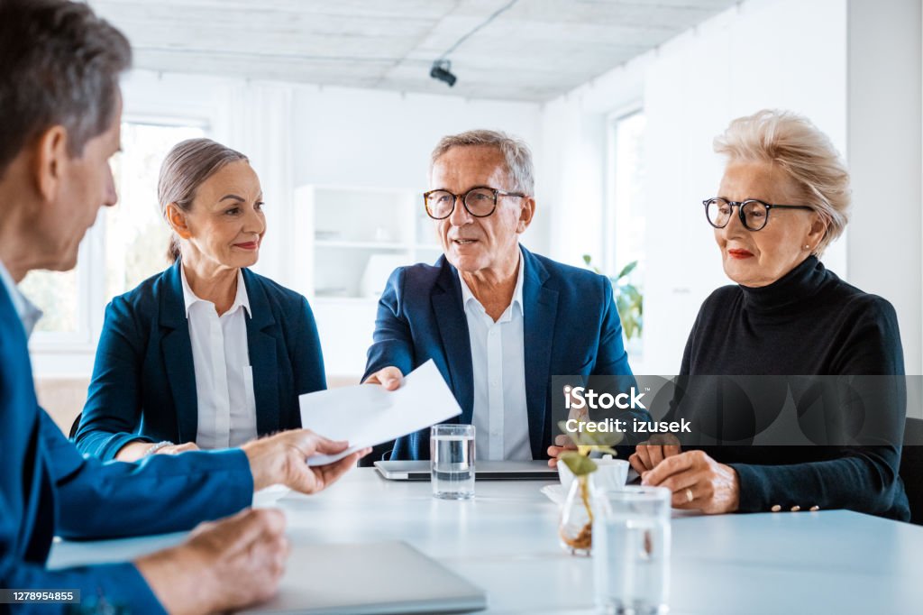 Senior business people during meeting Senior businesswomen and businessmen wearing elegant suits sitting at the table in the office and discussing during business meeting, exchanging contract. Corporate Governance Stock Photo