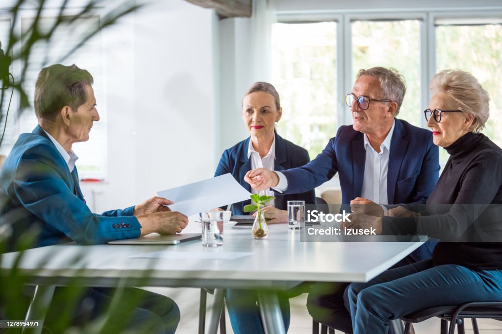 Senior business people during meeting Senior businesswomen and businessmen wearing elegant suits sitting at the table in the office and discussing during business meeting, exchanging contract. Business Meeting Stock Photo