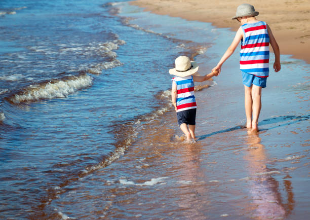 Two brothers barefoot are walking on the beach in water. Boys are on vacation in summer at the sea.