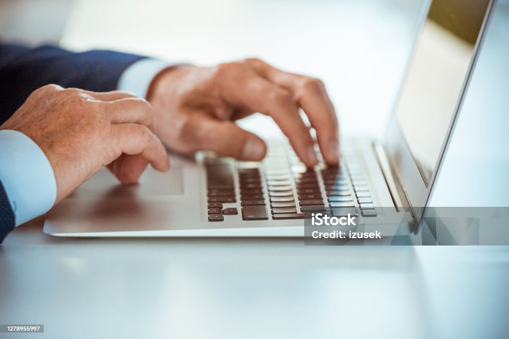 Senior businessmen using laptop Senior entrepreneur wearing elegant suits sitting at the table in the office, working on laptops. Close up on hands, unrecognizable person. Blue Stock Photo