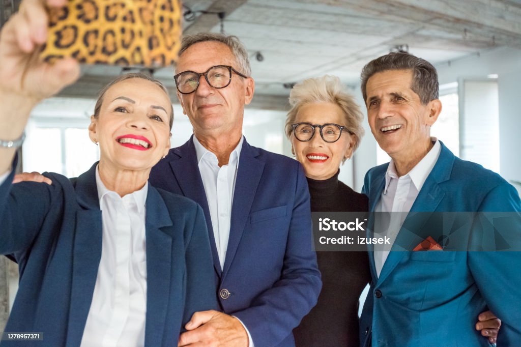 Successful business people taking selfie Cheerful senior businesswomen and businessmen wearing elegant suits standing in the modern office and talking selfie using smart phone. Senior Adult Stock Photo