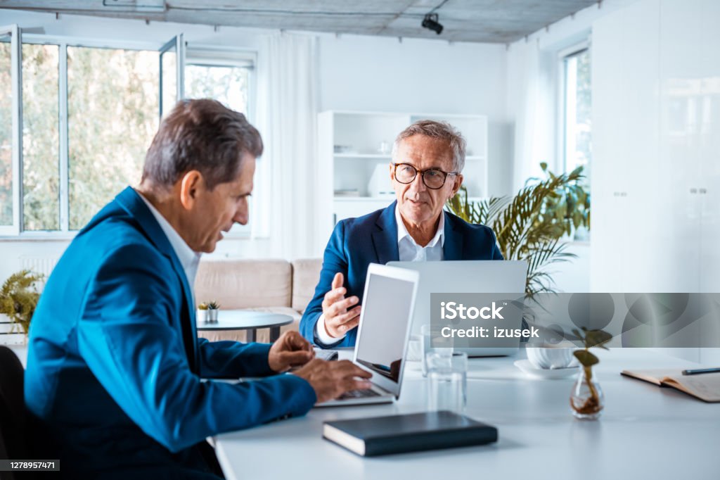 Two senior businessmen in the office Senior entrepreneurs wearing elegant suits sitting at the table in the office, working on laptops and discussing. Business Stock Photo