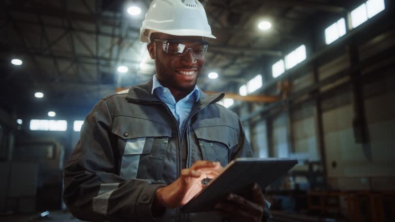 Professional Heavy Industry Engineer/Worker Wearing Safety Uniform and Hard Hat Uses Tablet Computer. Smiling African American Industrial Specialist Walking in a Metal Construction Manufacture.