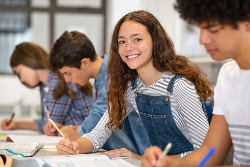 Satisfied young woman looking at camera while her friends studying at college. Team of multiethnic students preparing for university exam. Portrait of beautiful girl with freckles sitting in a row with her classmates during high school exam.