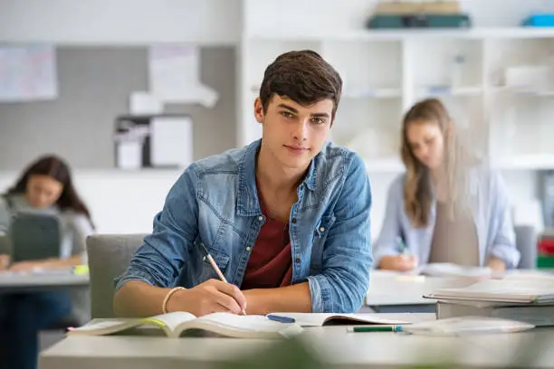 Happy student taking notes while studying in high school. Satisfied young man looking at camera while sitting at desk in classroom. Portrait of college guy writing while completing project and assignment.