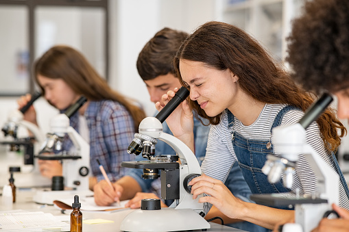 Group of multiethnic college students performing experiment using microscope in science lab. University focused student looking through microscope in biology class while sitting in a row at desk. High school girl examine samples during lecture.