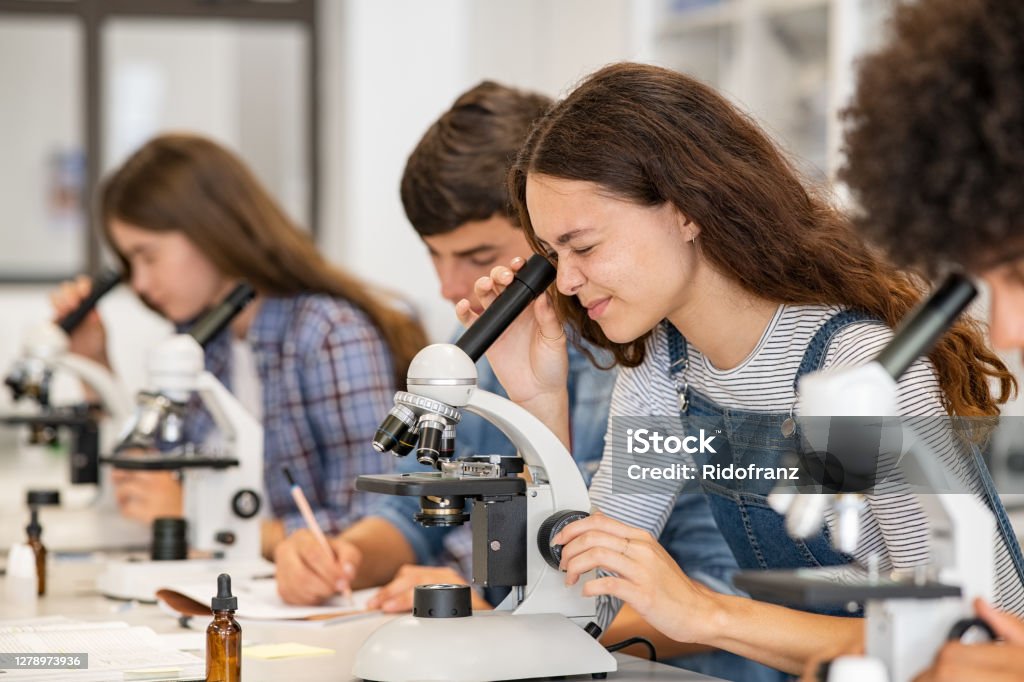 Biology class at high school lab Group of multiethnic college students performing experiment using microscope in science lab. University focused student looking through microscope in biology class while sitting in a row at desk. High school girl examine samples during lecture. Science Stock Photo