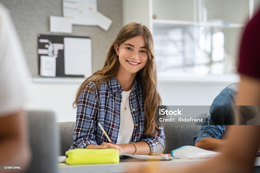 Smiling college girl studying in classroom Beautiful university student smiling while studying for exams in classroom. Pretty woman sitting in classroom full of students during class. Portrait of happy young woman writing notes and looking at camera. Teenager Stock Photo