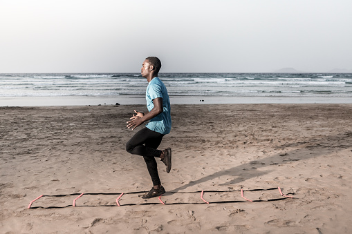 Side view of focused African American sportsman doing ladder drill near sea during fitness training on Famara Beach in Lanzarote, Spain
