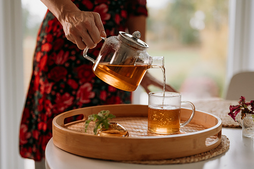 Woman tea ceremony pouring fresh brewed tea in glas cup and teapot\nPhoto taken indoors in dining room pn table