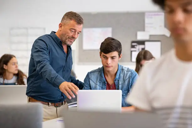 Professor assisting college student with laptop in classroom during computer lesson. Teacher talking and explaining something to guy sitting at desk. Mature man lecturer helping high school young man with laptop during lecture in university.