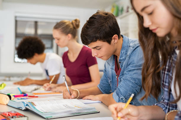 High school students doing exam in classroom Group of college students studying in classroom writing notes during lesson. Focused guy and girls studying in college library sitting at desk. Group of multiethnic university students doing research sitting in a row. adolescence stock pictures, royalty-free photos & images