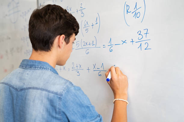 College student solving math equation on white board Back view of high school student solving math problem on whiteboard in classroom. Young man writing math solution on white board using marker. College guy solving math expression during lesson. mathematics stock pictures, royalty-free photos & images