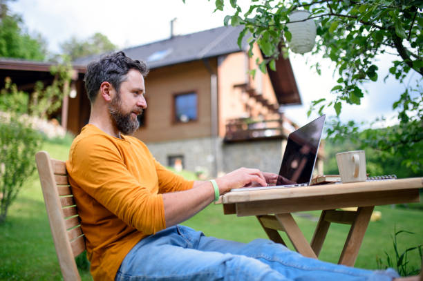 Side view of man with laptop working outdoors in garden, home office concept. Side view of mature man with laptop working outdoors in garden, home office concept. rural scene stock pictures, royalty-free photos & images