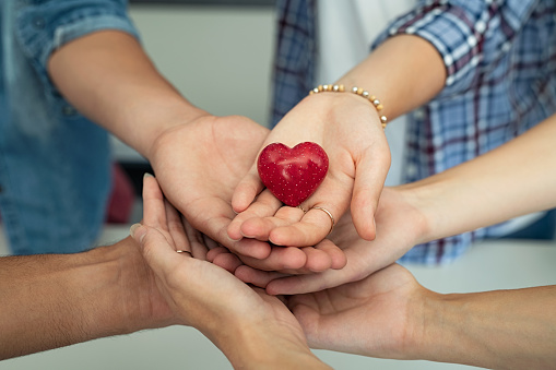 Close up of friends hands holding a red heart shape. Top view of young men and women hands showing small red heart. Multiethnic guys and girls holding a heart-shaped stone symbol of love and care.