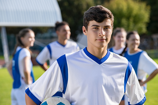 Portrait of young satisfied soccer player holding a football and looking at camera. Young man during training on soccer field with copy space. Proud teen guy training on soccer field with school teammmates in background.