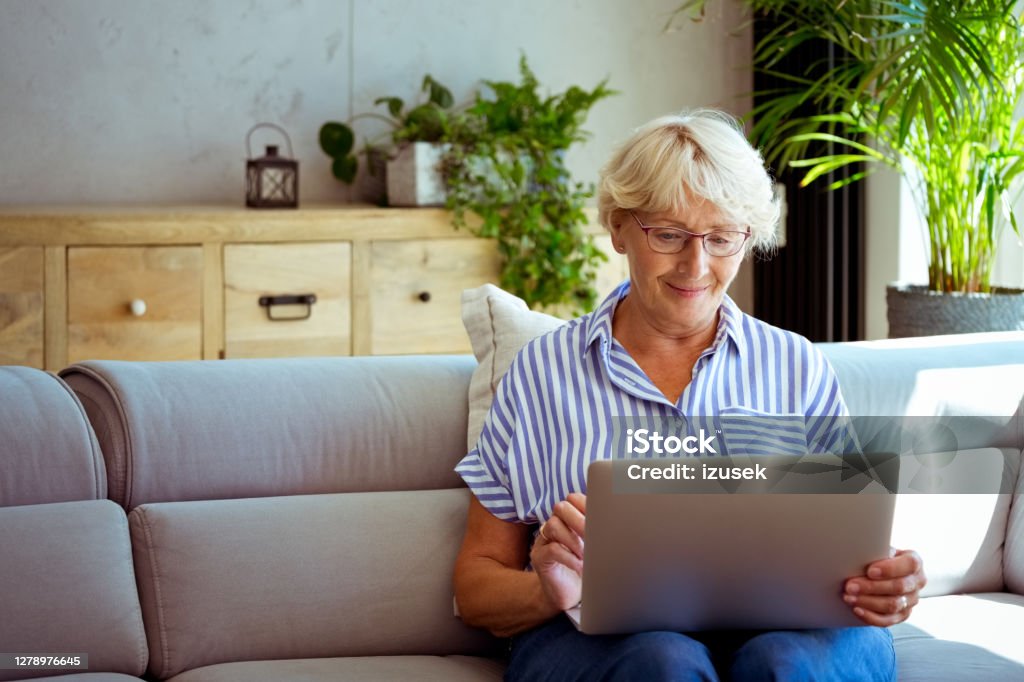 Senior woman using laptop at home Smiling elderly lady having video conference, using laptop, sitting on sofa in the living room at home. Domestic Life Stock Photo