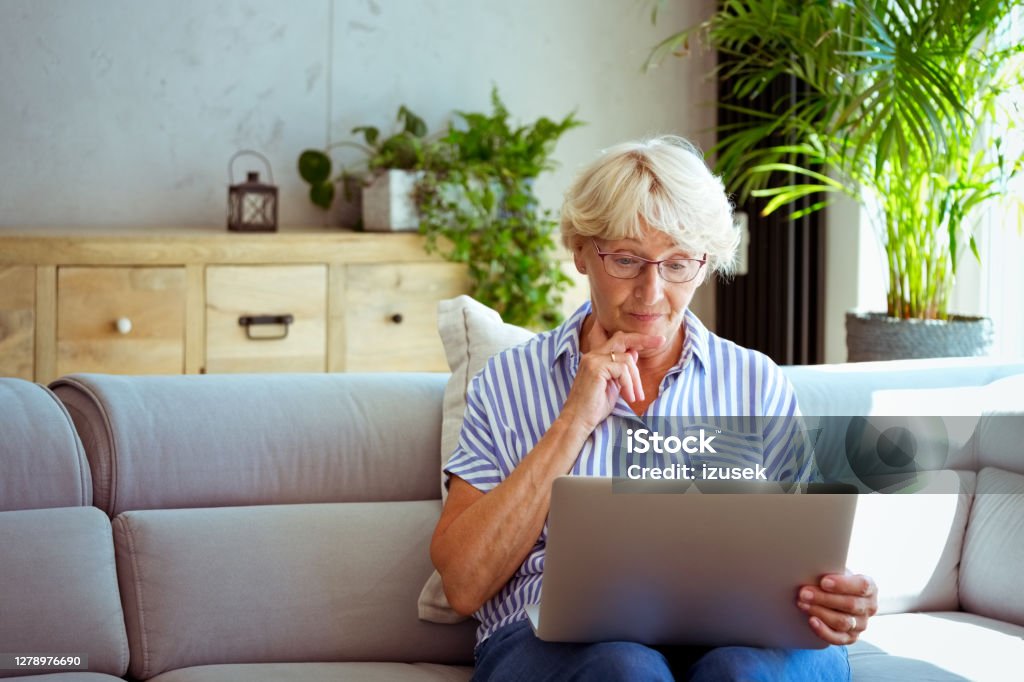 Senior woman using laptop at home Worried elderly lady having video conference, using laptop, sitting on sofa in the living room at home. Senior Adult Stock Photo