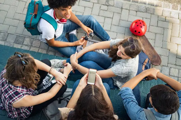 High angle view of guys and girls sitting and talking together. Cheerful group of happy friends in conversation after class. Top view of group of carefree teens spending time together after school while young woman using smart phone.