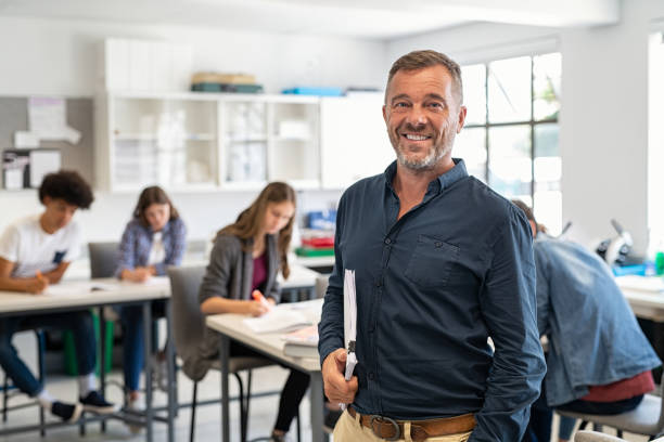 Mature man professor standing in class Portrait of mature teacher standing in university library and looking at camera with copy space. Happy mid adult lecturer at classroom standing after giving lecture. Satisfied high school teacher smiling and looking at camera while his studets studying in background. teacher stock pictures, royalty-free photos & images