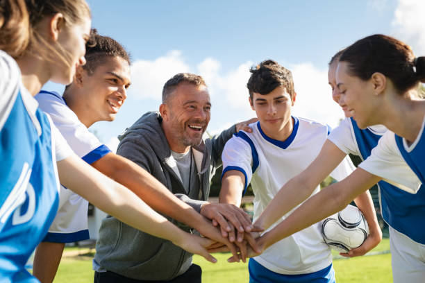 Team of young football players stacking hands before match Happy coach and high school soccer team with their hands stacked in football court. Young football players stacking hands together. Mature man coach encourages his students to do their best during the sport match. stacked hands photos stock pictures, royalty-free photos & images