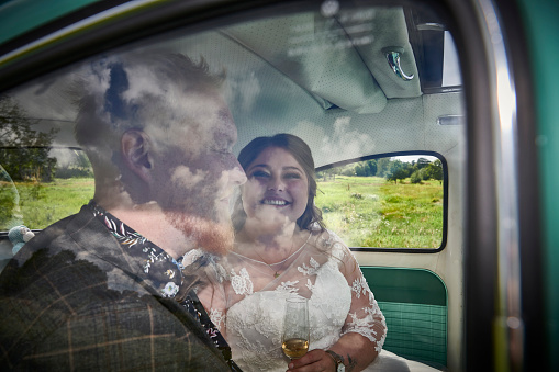 Photo shoot with newlywed young couple real people looking happy. Smiling sitting on the back seat in a green car. Reflects in the car window of the sky